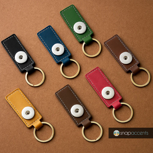 Leather snap keychains in black, blue, green, brown, red, and yellow with gold rings, featuring white snap buttons for interchangeable SnapAccents jewelry pieces, displayed on a brown background with SnapAccents logo.