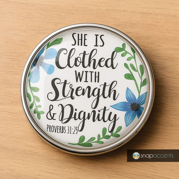 Round SnapAccents snap jewelry charm with inspirational Proverbs 31:25 quote "She is clothed with strength & dignity," decorated with blue flowers and green leaves on white background, silver metal frame, placed on wooden surface.