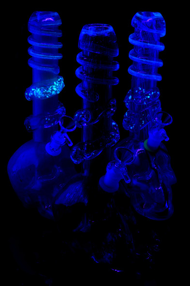Soft-glass skull beaker water pipes under black light showing glow accent ring and spiral wrap; retail head-shop merchandising detail on dark backdrop.