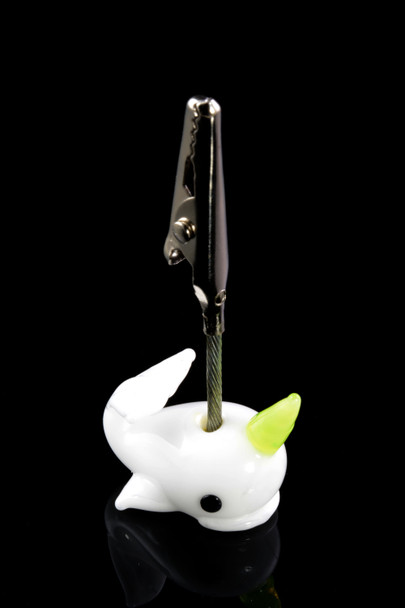 White glass narwhal with lime horn holding a metal alligator clip on braided stem, glossy black backdrop—wholesale roach clip accessory, case-ready.
