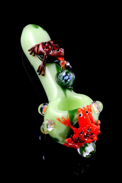 Angled rear three-quarter view of small frog-themed glass spoon for retail smoke gear, light-green body with sculpted red and brown frogs on black.