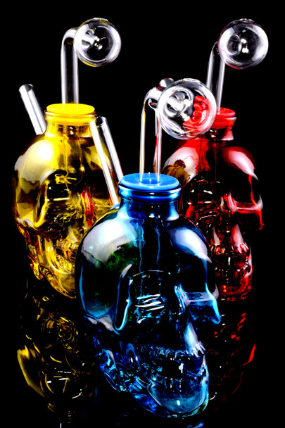 Close trio of 4" glass skull oil burner rigs on black with clear domes—head-shop wholesale detail in red, yellow, and blue.