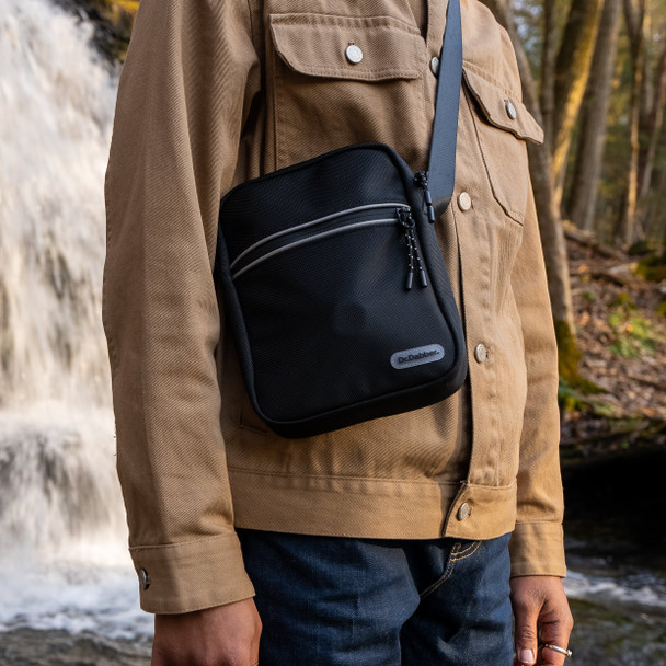 Black Dr. Dabber tour bag worn crossbody outdoors, lifestyle shot showing textured shell, zipper pocket, and wide strap for head-shop displays.