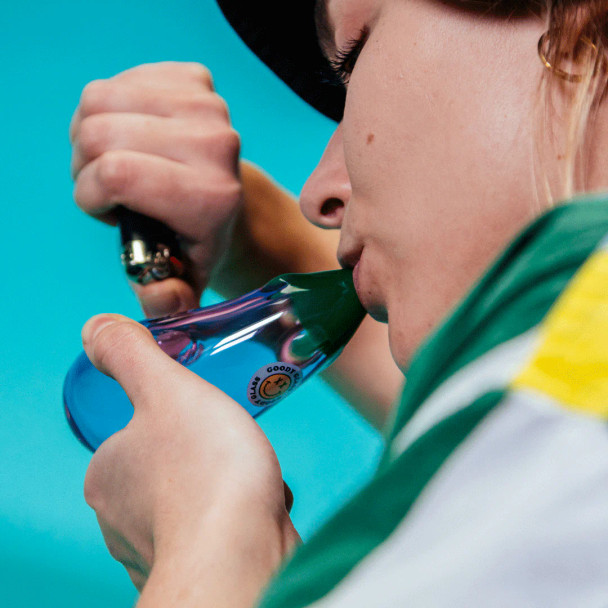 Person holding Goody Glass eggplant hand pipe with purple body and green mouthpiece against teal backdrop