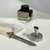 A feather pen set features a black ink bottle on a clear acrylic stand, a white feather quill with an ornate silver handle, and a matching silver pen holder with intricate designs, all resting on a white surface.