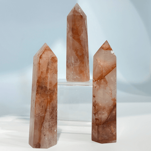 Three tall, pointed, polished crystal towers, identified as Red Fire Quartz, showcasing clear quartz with vibrant reddish-orange inclusions that resemble flames. Two are in the foreground, and one is elevated on a clear stand in the background. Three tall, pointed, polished crystal towers, identified as Red Fire Quartz, showcasing clear quartz with vibrant reddish-orange inclusions that resemble flames. Two are in the foreground, and one is elevated on a clear stand in the background.