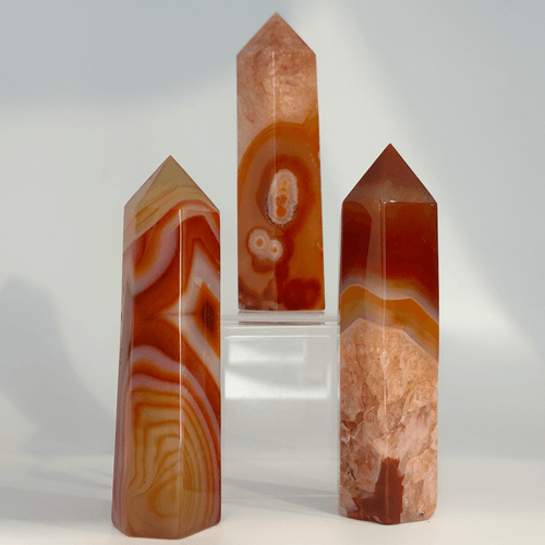 A trio of Carnelian crystal towers arranged at different heights. One point is elevated on a clear acrylic display stand, showcasing its intricate, circular agate-like patterns in warm shades of amber and terracotta. The smooth, faceted edges of these polished gemstone towers create a modern, geometric silhouette perfect for meditation or home decor.