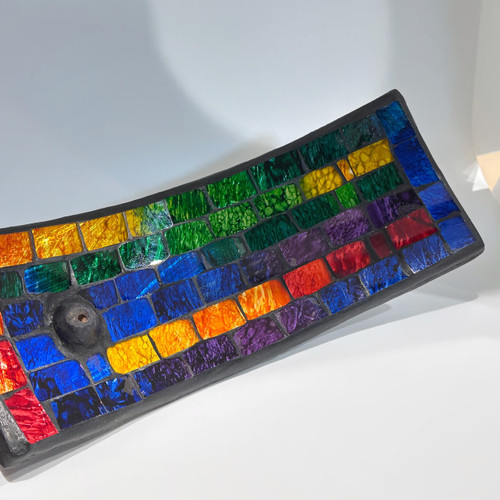 A close-up of a dark, rectangular incense tray with a rainbow mosaic of small, square tiles. It features a circular indentation for an incense stick and curves upward slightly.