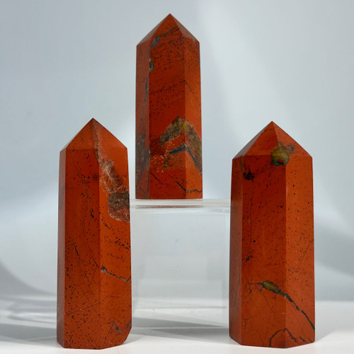 A trio of polished Red Jasper crystal points are arranged on a clear display stand. The stones are a deep, mottled red with hints of brown. A trio of polished Red Jasper crystal points are arranged on a clear display stand. The stones are a deep, mottled red with hints of brown.