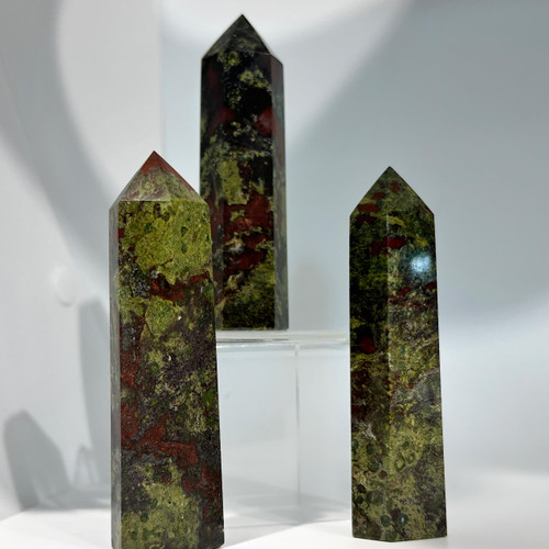 A trio of polished crystal points, mottled in dark green and deep reddish-brown colors, are arranged on a clear display stand against a white background. A trio of polished crystal points, mottled in dark green and deep reddish-brown colors, are arranged on a clear display stand against a white background.