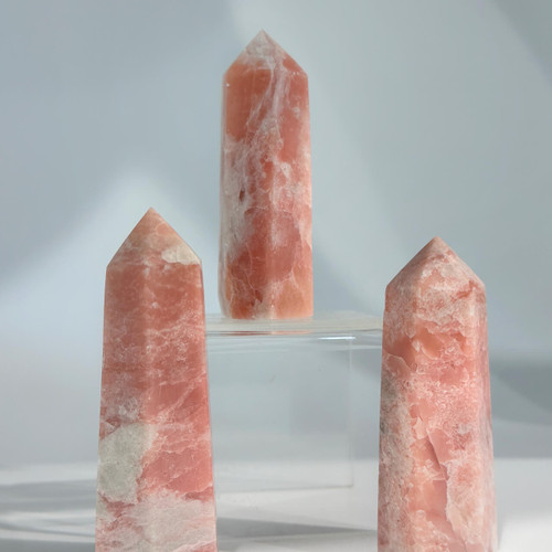 A trio of polished Pink Opal crystal points, with soft pink and white swirls, are arranged on a clear display stand. A trio of polished Pink Opal crystal points, with soft pink and white swirls, are arranged on a clear display stand.