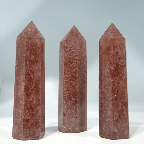 Three polished crystal towers, likely strawberry quartz, displaying a translucent reddish-pink color with tiny white inclusions, set against a light background. Three polished crystal towers, likely strawberry quartz, displaying a translucent reddish-pink color with tiny white inclusions, set against a light background.
