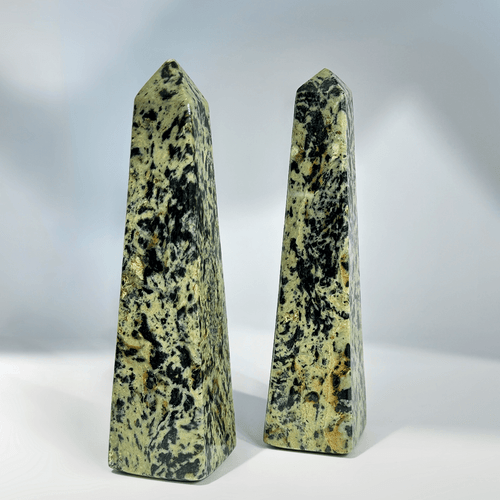 A pair of tall, pointed crystal towers (obelisks) made of Dragi "Dragon" Jade (nephrite). The stones are primarily mottled olive green and black with hints of yellow/brown, and they are displayed against a white background.