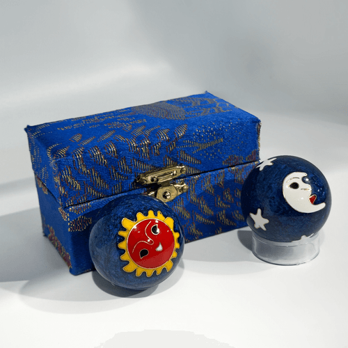 Two deep blue, marble-textured meditation spheres displayed next to their matching storage box. One sphere features a vibrant red sun with a yellow border and a stylized face, while the other shows a white crescent moon with a face and small white stars. Behind them sits a rectangular, blue silk-covered box with a gold-toned metal latch and intricate woven patterns. The entire set is presented on a plain white surface.