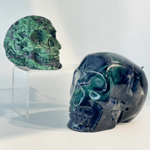 On the right, in the foreground, is the glossy Rainbow Fluorite skull, carved with smooth, polished details that reveal deep bands of translucent teal, purple, and dark blue. On the left, elevated in the background on a clear acrylic riser, is the Ruby Zoisite skull, shown in profile to highlight its intricate carving and natural, matte blend of green zoisite, black speckles, and purple-red ruby deposits.