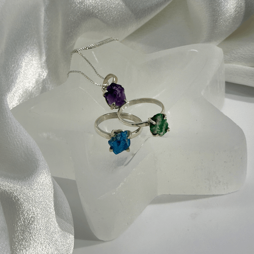 A star-shaped jewelry dish carved from translucent white selenite, holding two silver rings with raw blue and green gemstones and a purple amethyst pendant necklace.