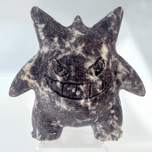 A gemstone carving of the Pokemon Gengar crafted from Purple Mica. The stone has a deep plum and lavender base with swirling white and grey mineral inclusions, giving it a marbled, shimmering appearance. The figurine captures Gengar’s iconic stout body, pointed ears, spiked back, and a large, carved toothy smile.