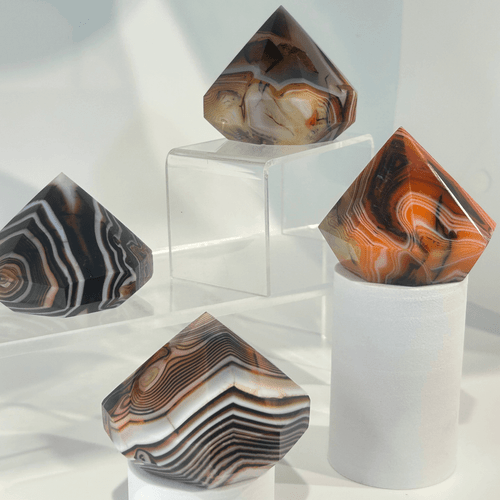 Four polished sardonyx gemstones carved into diamond shapes, shown in a close-up arrangement. The patterns range from thin, zebra-like black and white stripes to broad, vibrant orange and tan swirls.