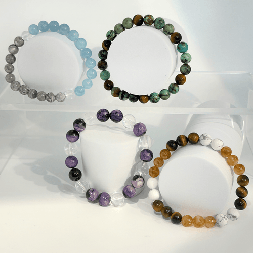 A clear, four-pedestal display on a white background showcasing all four gemstone beaded element bracelets. The set includes a light blue/grey water bracelet, a green/brown earth bracelet, a purple/clear air bracelet, and a multi-colored brown/white/orange fire bracelet.