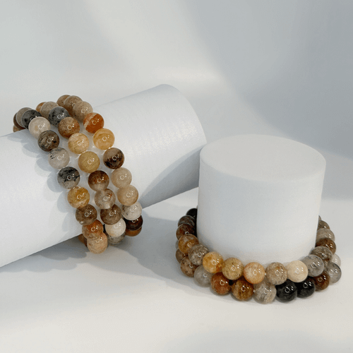 Garden Quartz gemstone bead bracelets displayed on white cylindrical stands, featuring polished natural stone beads in earthy brown, gray, amber, and cream tones.