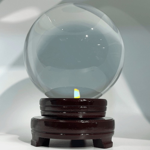 Clear crystal ball on a polished dark brown wooden base, illuminated from below, with a softly blurred light gradient background.