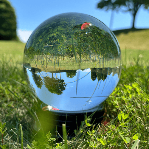 Crystal ball resting on green grass, reflecting the blue sky and surrounding trees, with sunlight casting highlights on its surface.