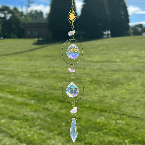 Hanging sun catcher with a gold sun charm, purple amethyst stones, iridescent faceted crystals, and a pointed blue pendant hanging outdoors.