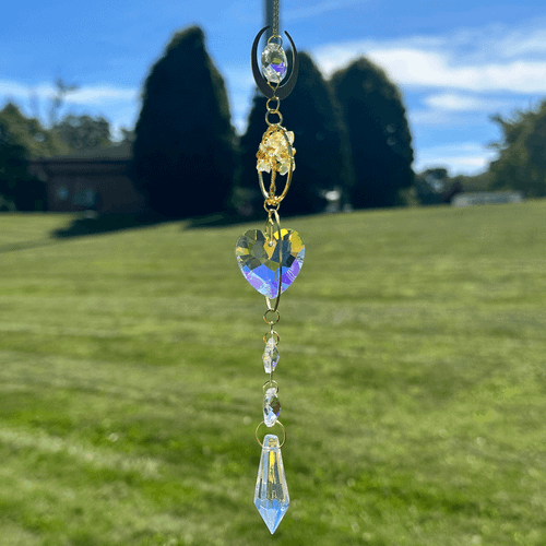 Gold-toned hanging crystal suncatcher with yellow stones, a heart-shaped rainbow-reflective crystal, and pointed prism against a blurred lawn and blue sky.