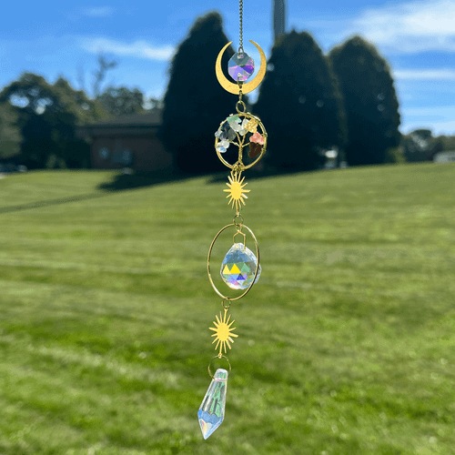 Hanging chakra tree of life suncatcher with colorful stones, gold crescent and sunburst charms, and faceted crystals against a blurred grassy outdoor background.