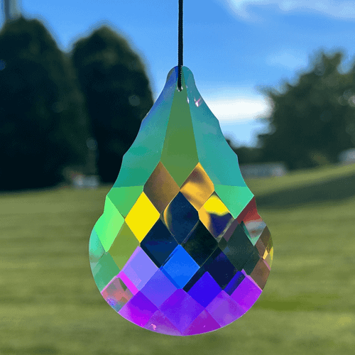 Faceted multicolored teardrop suncatcher hanging outdoors against a blurred blue sky, trees, and grassy field.