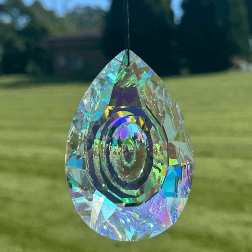 Faceted teardrop sun catcher with a reflective indented oval center hanging against a blurred green lawn and trees.