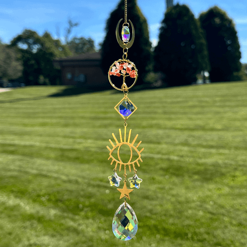 Gold-toned Tree of Life suncatcher with orange-red stones, crystals, sun and star charms, hanging outdoors against a blurred lawn and trees.