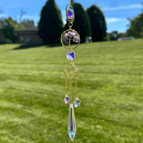 Gold tree of life suncatcher with purple amethyst stones, faceted crystals, and long iridescent prism against a blurred grassy outdoor background.
