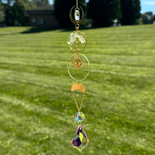 Gold-tone tree of life suncatcher with pale green stones, a sun-face charm, and colorful faceted crystals hanging outdoors.