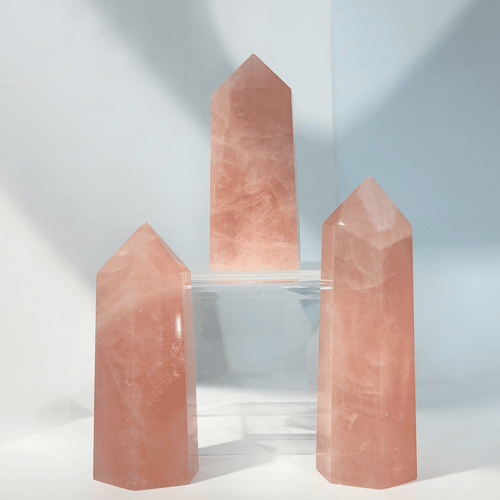 Three natural Rose Quartz crystal points, faceted and polished into hexagonal towers. The soft pink stones show natural internal cloudy inclusions and are displayed on clear risers against a light backdrop. Three natural Rose Quartz crystal points, faceted and polished into hexagonal towers. The soft pink stones show natural internal cloudy inclusions and are displayed on clear risers against a light backdrop.