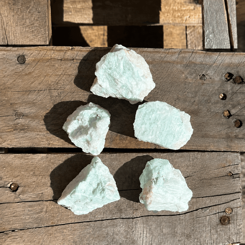 Rough Amazonite stones with pale blue-green coloring displayed on a rustic wooden surface, showcasing their natural, unpolished texture.