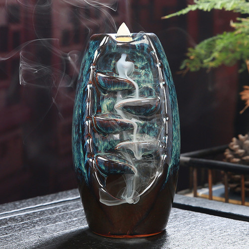 A brown and blue drip leaf backflow burner with swirling white smoke, displaying carved leaves and a glossy finish, set against a blurred natural backdrop.