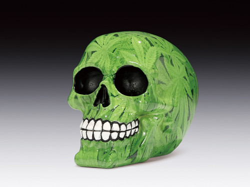 Green decorative skull with black eye sockets and white teeth, adorned with painted cannabis leaves, set against a dark background.