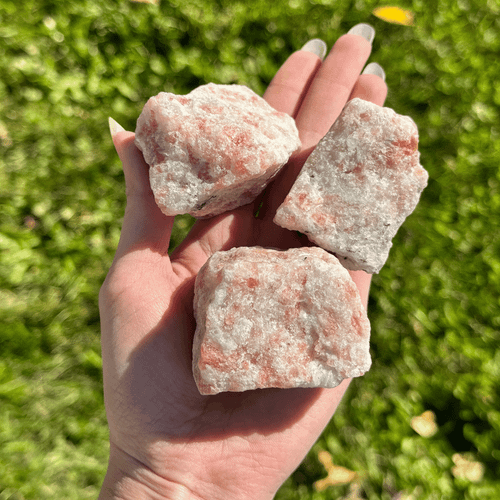 Rough Golden Sunstone mineral specimens with pale peach, pink, and cream-colored textures displayed in a person's hand against a green grassy background.