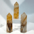 A set of three tall, polished Golden Healer Quartz tower points, displaying clear and smoky quartz with prominent internal layers of golden-yellow and brown Hematoid mineral inclusions, displayed on a clear stand. A set of three tall, polished Golden Healer Quartz tower points, displaying clear and smoky quartz with prominent internal layers of golden-yellow and brown Hematoid mineral inclusions, displayed on a clear stand.