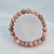 A top-down, central view of a single natural pink opal beaded stretch bracelet. The highly polished gemstone beads exhibit a uniform size with rich natural variations in color, ranging from creamy blush pink to deeper rose and mauve-grey inclusions. The smooth texture of the beads contrasts cleanly with the matte white background.