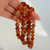 A close-up view of three 8mm polished Red Agate gemstone bracelets being held up by a hand against a soft white background. The bracelets drape vertically, catching the light to highlight the natural variety of the stones. The glossy beads feature a stunning mix of fiery orange, translucent amber, and deep rust-red hues, with several beads prominently displaying intricate, wavy white and cream banding patterns that make each bracelet unique.