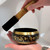 Tibetan bronze singing bowl with gold script, held in cupped hands, with a mallet poised above.