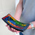 A curved, black incense holder features a vibrant mosaic of small, square tiles in rainbow colors. A thin, brown incense stick stands upright in a dark, circular indentation on the left side of the holder.