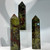 A trio of polished crystal points, mottled in dark green and deep reddish-brown colors, are arranged on a clear display stand against a white background.