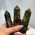 A hand holds three polished Dragon's Blood Jasper crystal points. The stones are mottled in colors of dark green and deep reddish-brown and are faceted to a point.