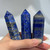 A hand holds three polished lapis lazuli crystal points. The stones are a deep blue with marbled patterns of lighter blue, grey, and sparkling flecks of gold pyrite.