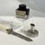 A feather pen set features a black ink bottle on a clear acrylic stand, a white feather quill with an ornate silver handle, and a matching silver pen holder with intricate designs, all resting on a white surface. A feather pen set features a black ink bottle on a clear acrylic stand, a white feather quill with an ornate silver handle, and a matching silver pen holder with intricate designs, all resting on a white surface.