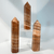 Three polished, pointed hexagonal stone obelisks made of Red Banded Calcite. The calcite features alternating horizontal bands of cream, white, and various shades of reddish-orange to rust brown. Three polished, pointed hexagonal stone obelisks made of Red Banded Calcite. The calcite features alternating horizontal bands of cream, white, and various shades of reddish-orange to rust brown.