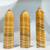 Three polished, pointed hexagonal stone obelisks made of Orange Calcite. The calcite features alternating horizontal bands of cream, white, and various shades of golden orange to honey yellow. Three polished, pointed hexagonal stone obelisks made of Orange Calcite. The calcite features alternating horizontal bands of cream, white, and various shades of golden orange to honey yellow.